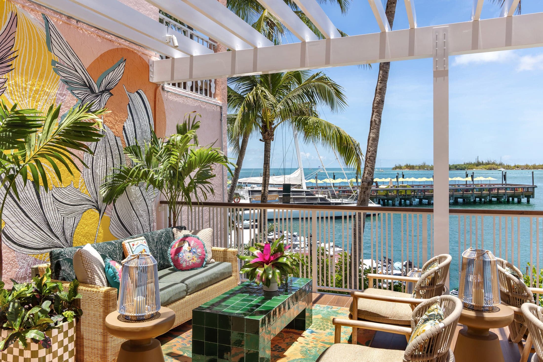 Covered waterfront terrace at Ocean Key Resort in Key West, Florida, with lounge seating, a green tile coffee table, tropical mural wall, and lantern accents, overlooking palm trees, the marina, and the water beyond.