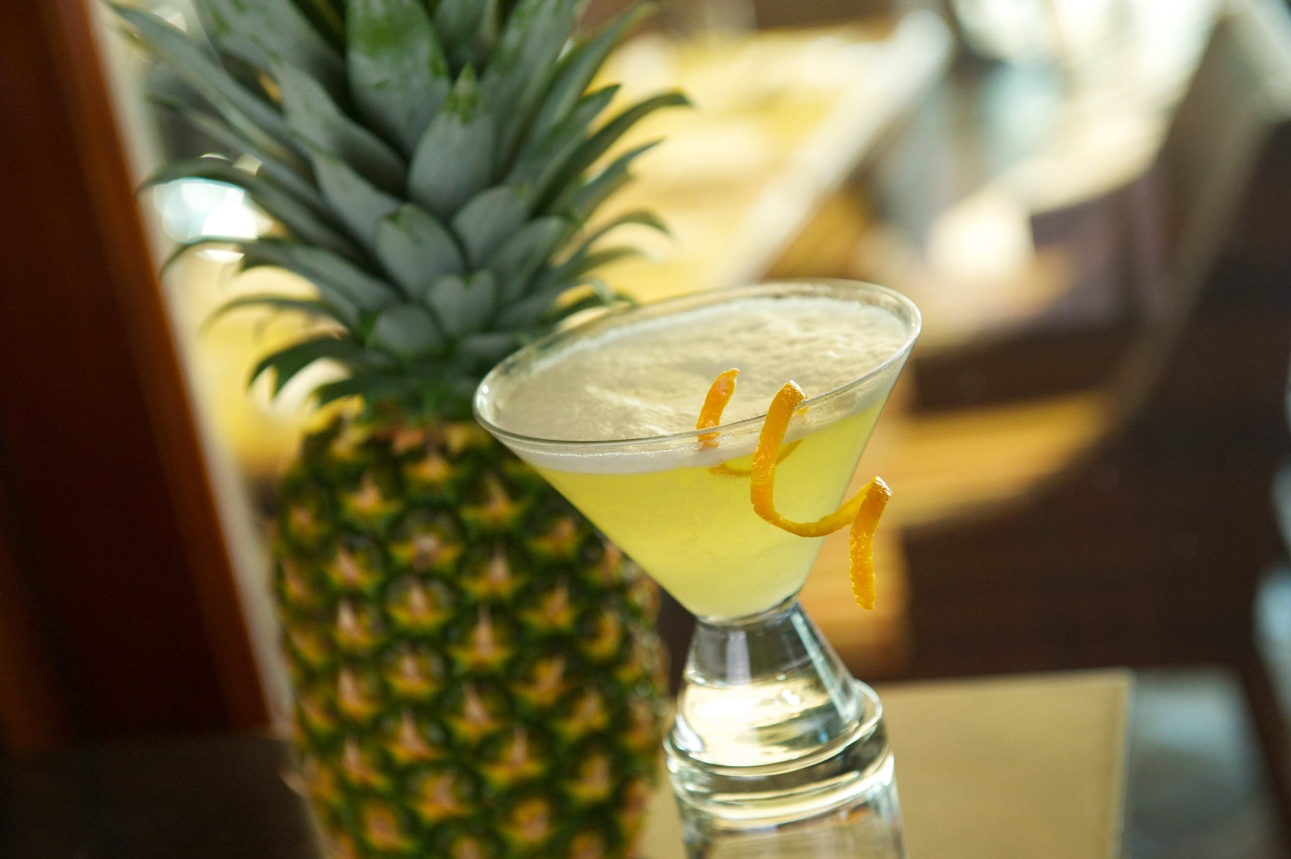 Frothy yellow cocktail in a martini glass with an orange peel garnish, next to a whole pineapple.