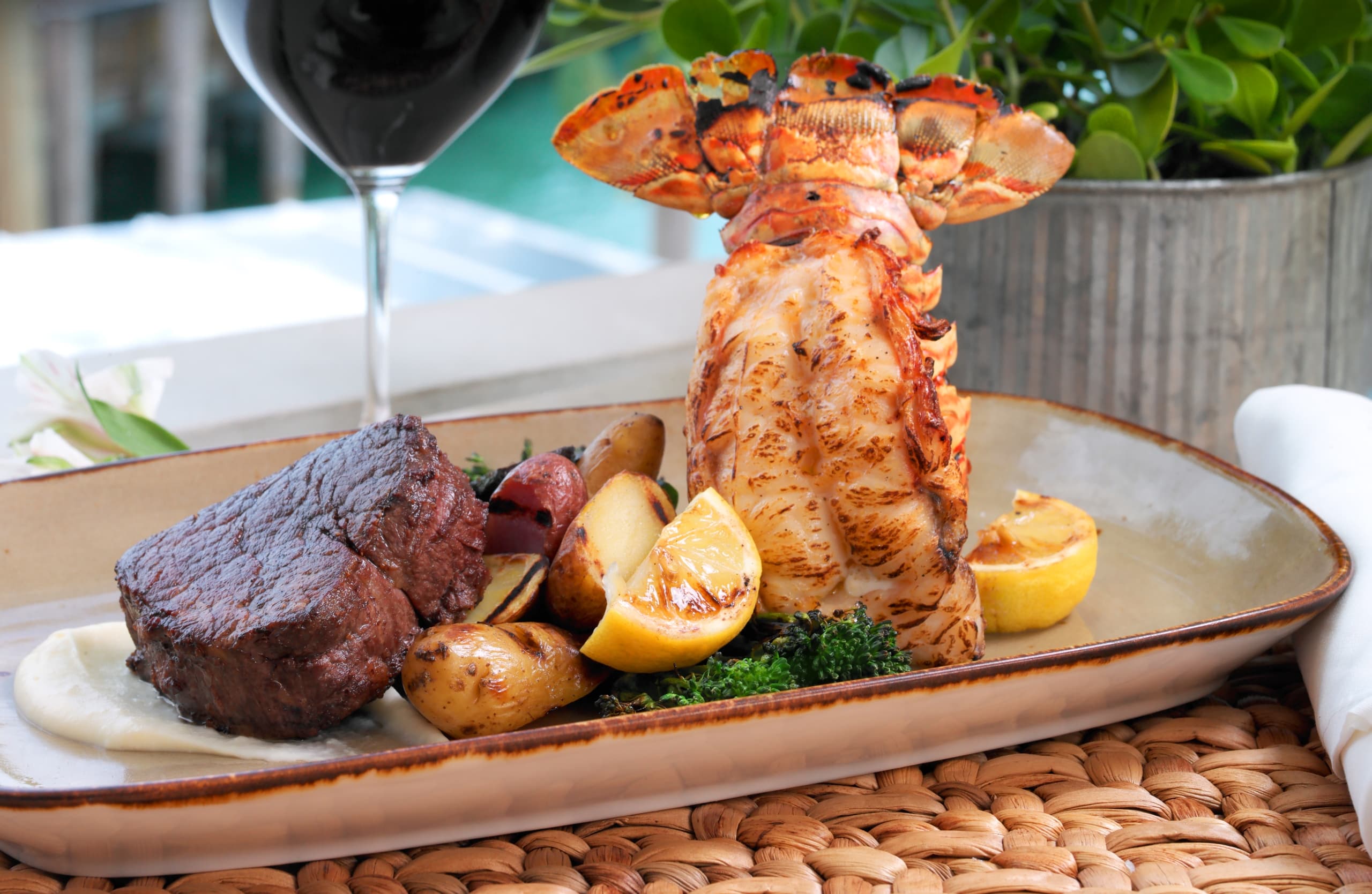Grilled filet mignon and lobster tail with roasted potatoes, broccolini, and lemon wedges over a white sauce, paired with red wine.