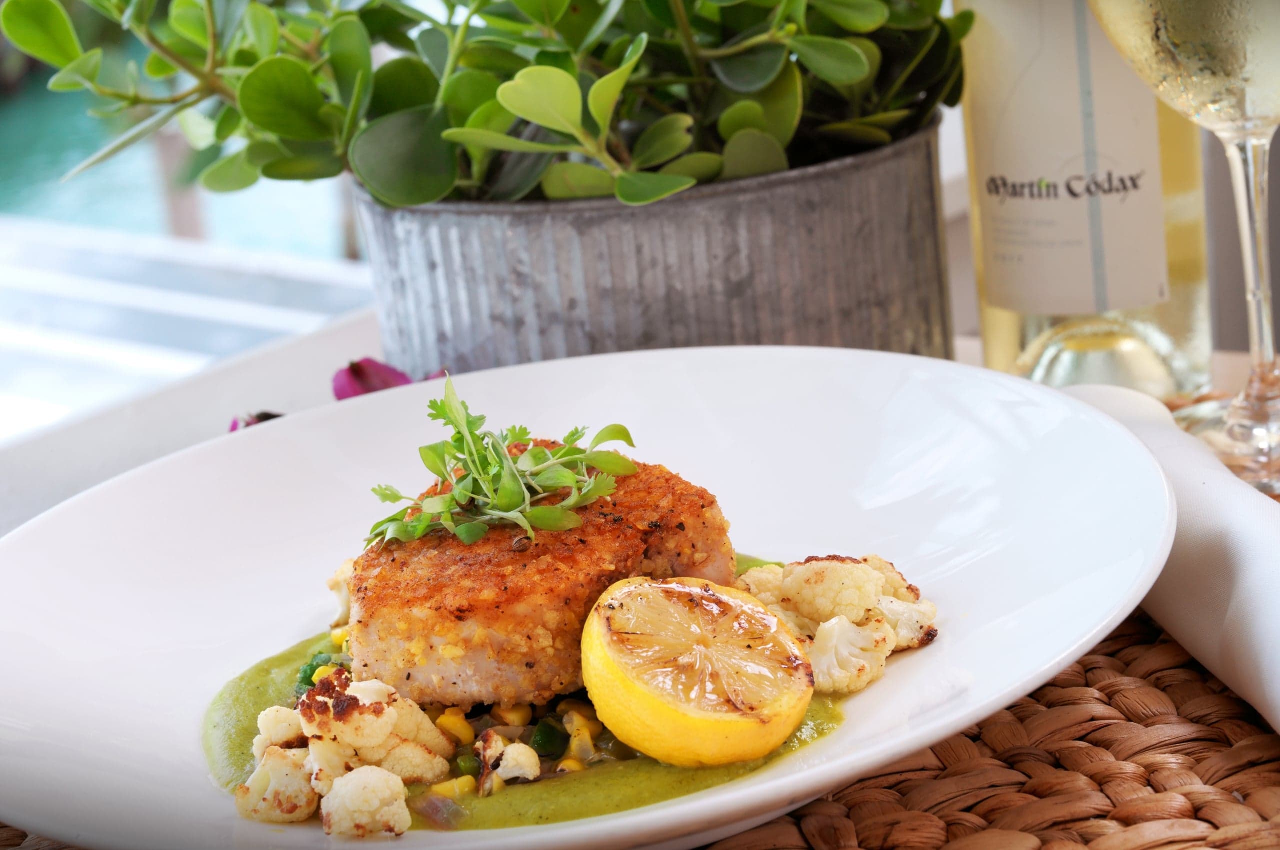 Panko-crusted fish fillet over green sauce with roasted cauliflower and a grilled lemon half, garnished with microgreens.