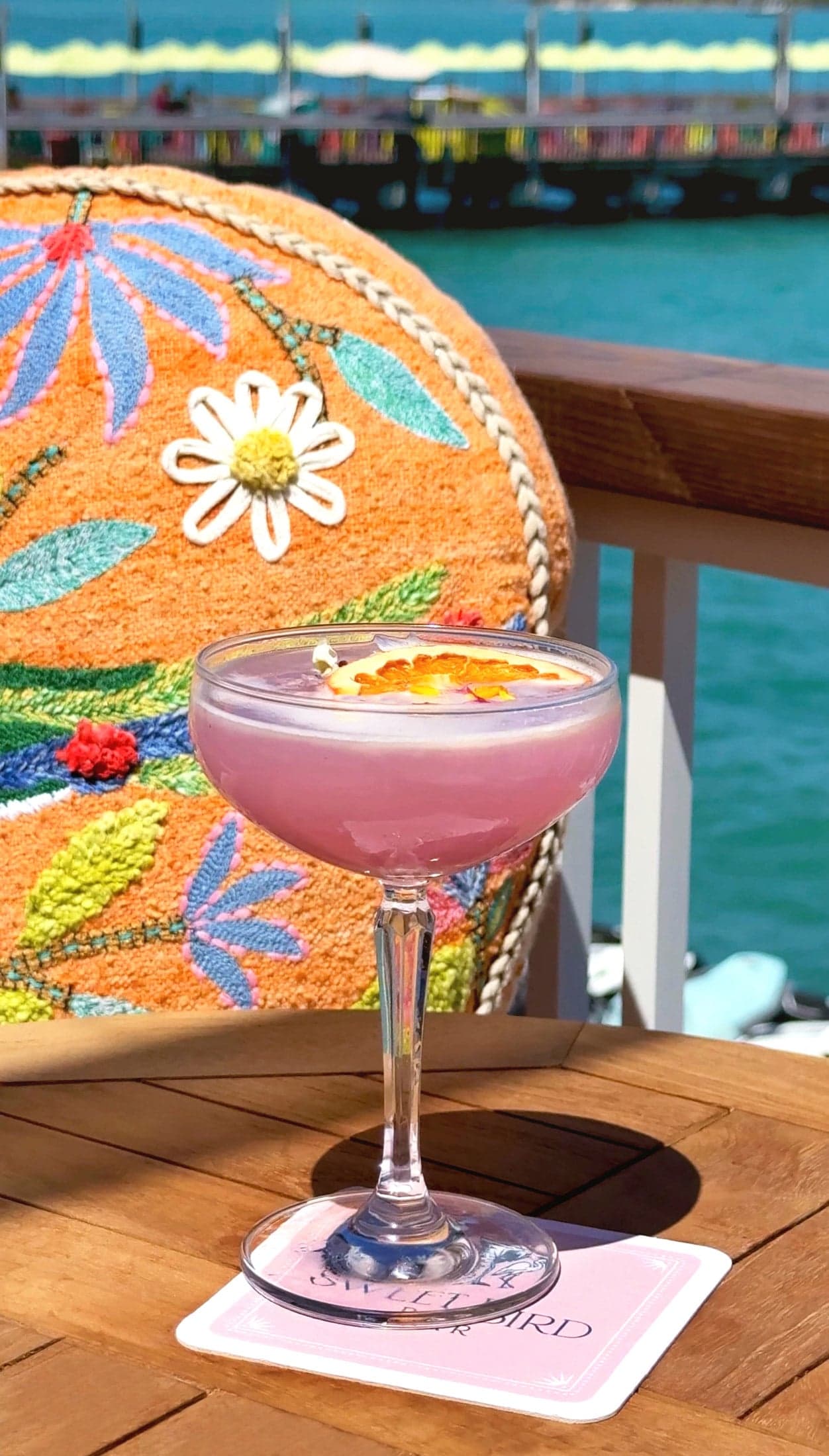 A pink craft cocktail in a coupe glass garnished with a dried citrus wheel, served on a wooden table at Ocean Key Resort in Key West, Florida, with a floral embroidered chair and waterfront marina view in the background.