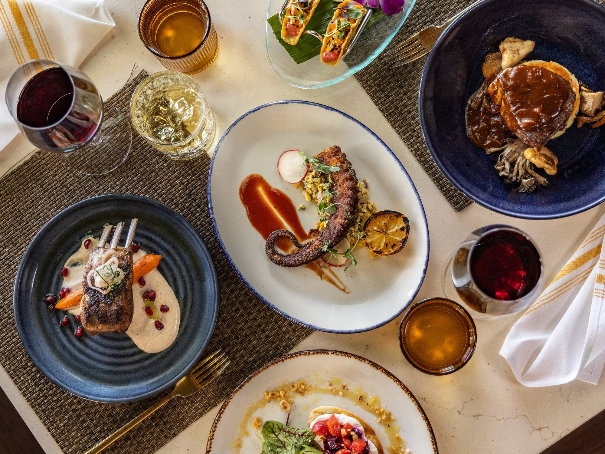 Overhead view of a dinner spread at Hot Tin Roof with grilled octopus, rack of lamb, filet mignon, tuna tacos, and wine