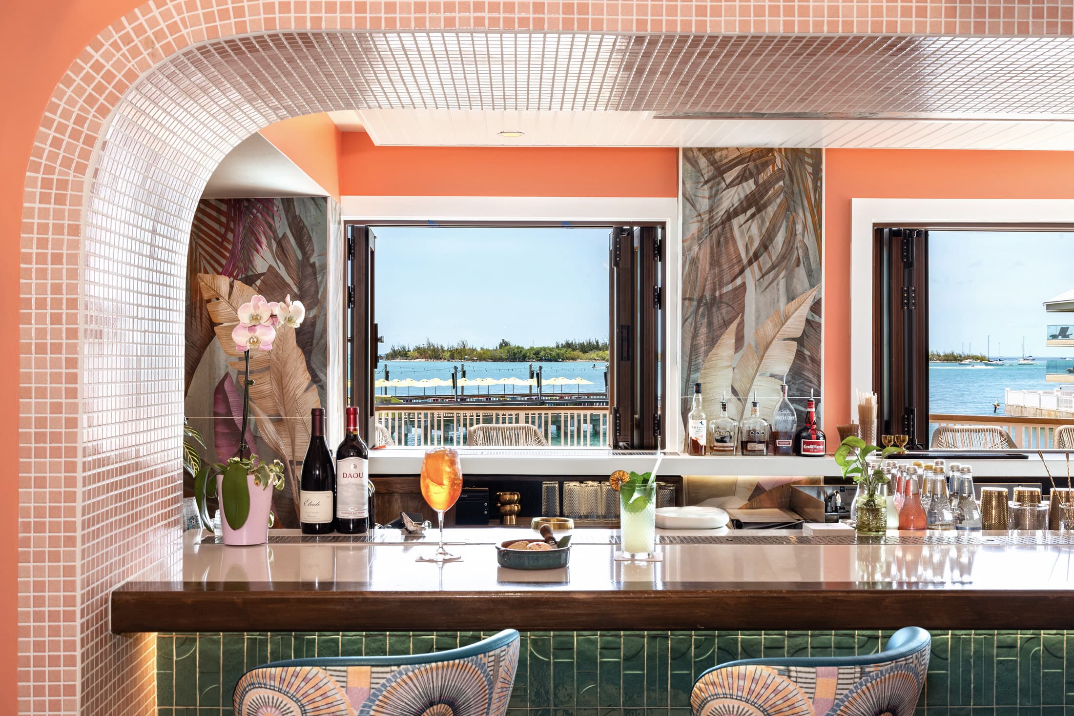 Waterfront bar at Sweet Bird Bar, Ocean Key Resort in Key West, Florida, with a pink tile archway framing the marble bar top, cocktails and wine bottles on display, patterned barstools, and open windows overlooking the marina and open water.